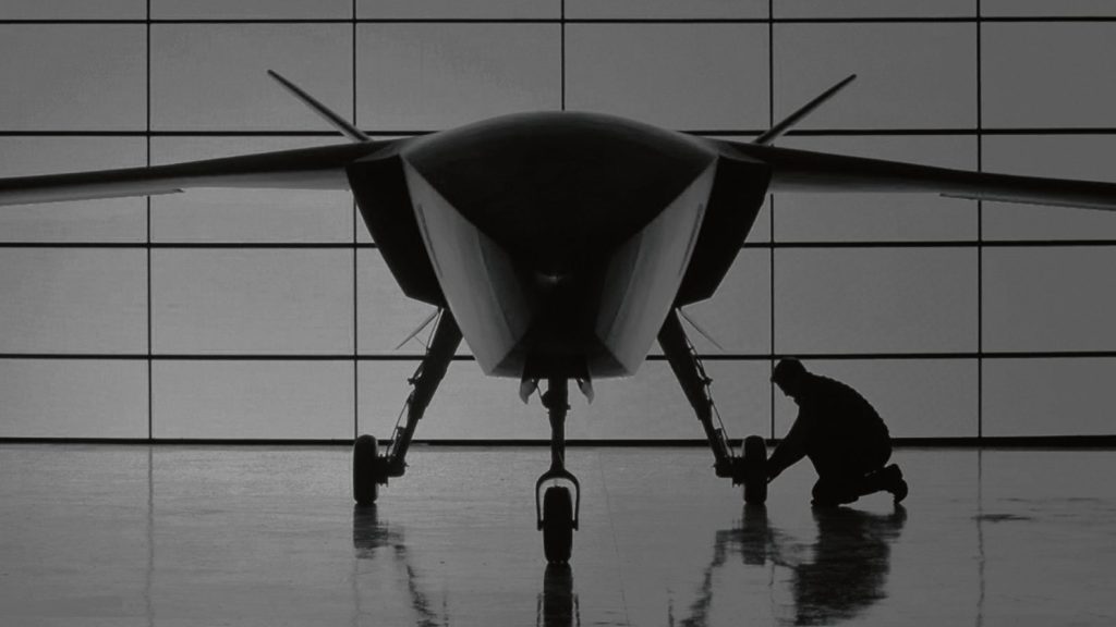 A large uncrewed combat aircraft in a hangar with a technician working at the nose wheel
