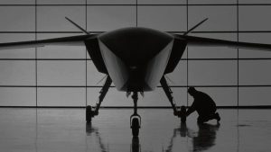 A large uncrewed combat aircraft in a hangar with a technician working at the nose wheel