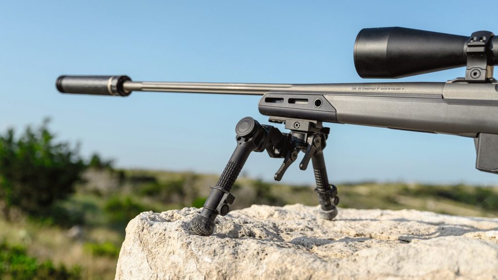 A short-barrelled hunting rifle with a sound moderator fitted, resting on a bipod on a rock